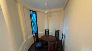 Un coin repas avec une table ronde et six chaises en bois, un ventilateur de plafond et une fenêtre agrémentée de rideaux bleus.