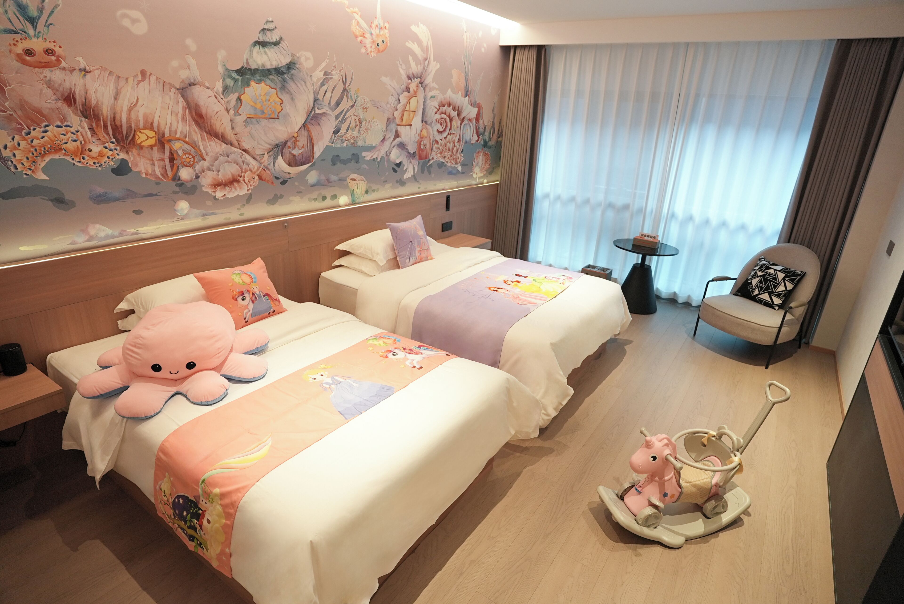 A hotel room with two beds, a mural on the wall, a rocking horse, and a chair.
