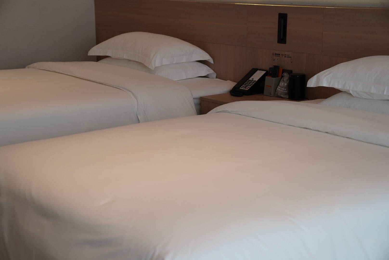 A hotel room with two beds, each with white bedding and pillows. There is a bedside table with a remote control, a phone, and a small storage box. The room has a wooden headboard and a wall-mounted light fixture.