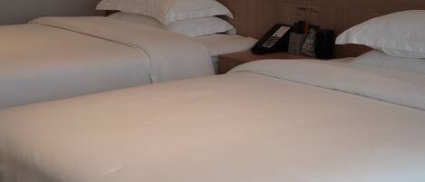 Habitación de hotel con dos camas, cada una con ropa de cama y almohadas blancas. Se observa una mesita de noche con un control remoto, un teléfono y un pequeño recipiente de almacenamiento. La habitación tiene un cabecero de madera y una lámpara de pared.