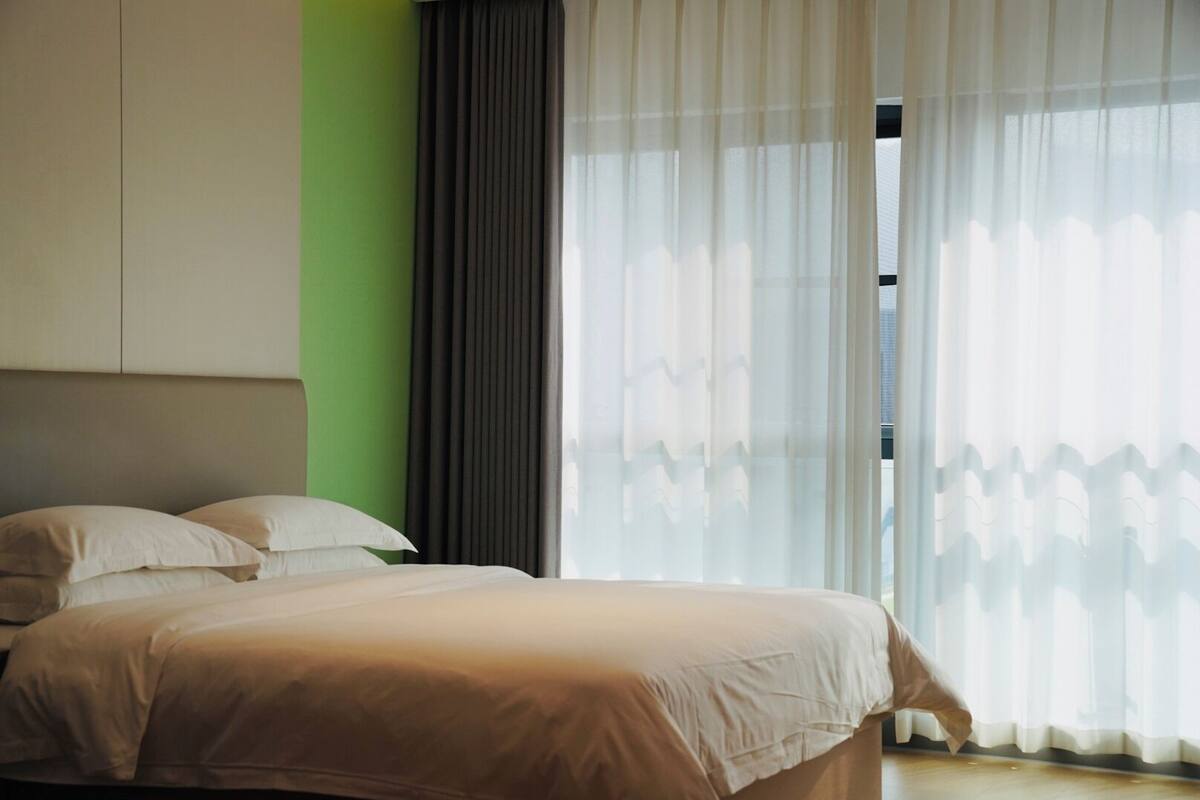 Ein Schlafzimmer mit Bett, Kopfstütze und Fenster mit durchsichtigen Vorhängen.