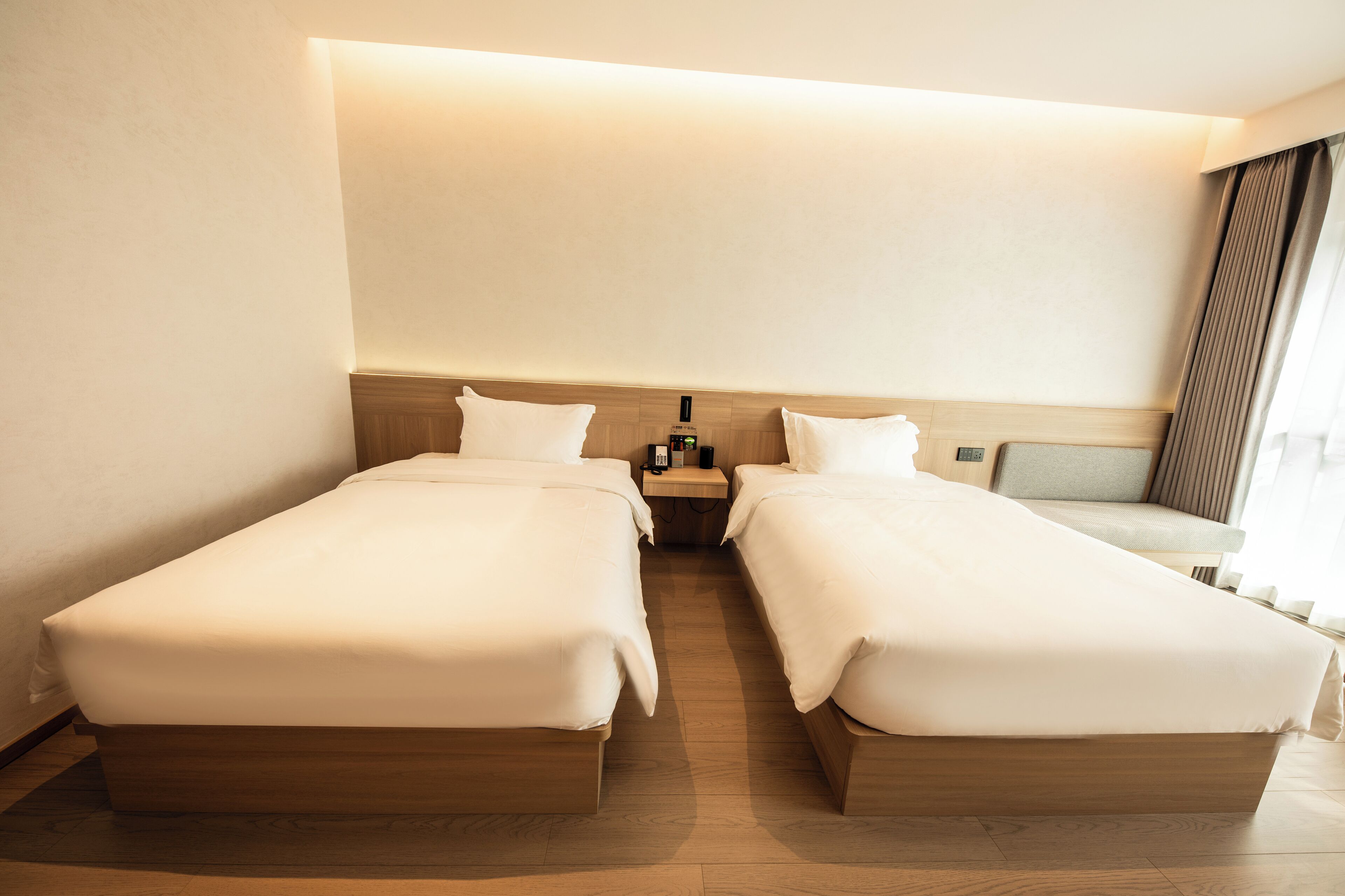 A hotel room with two single beds, a nightstand with a lamp, and a view of the outside through a window.