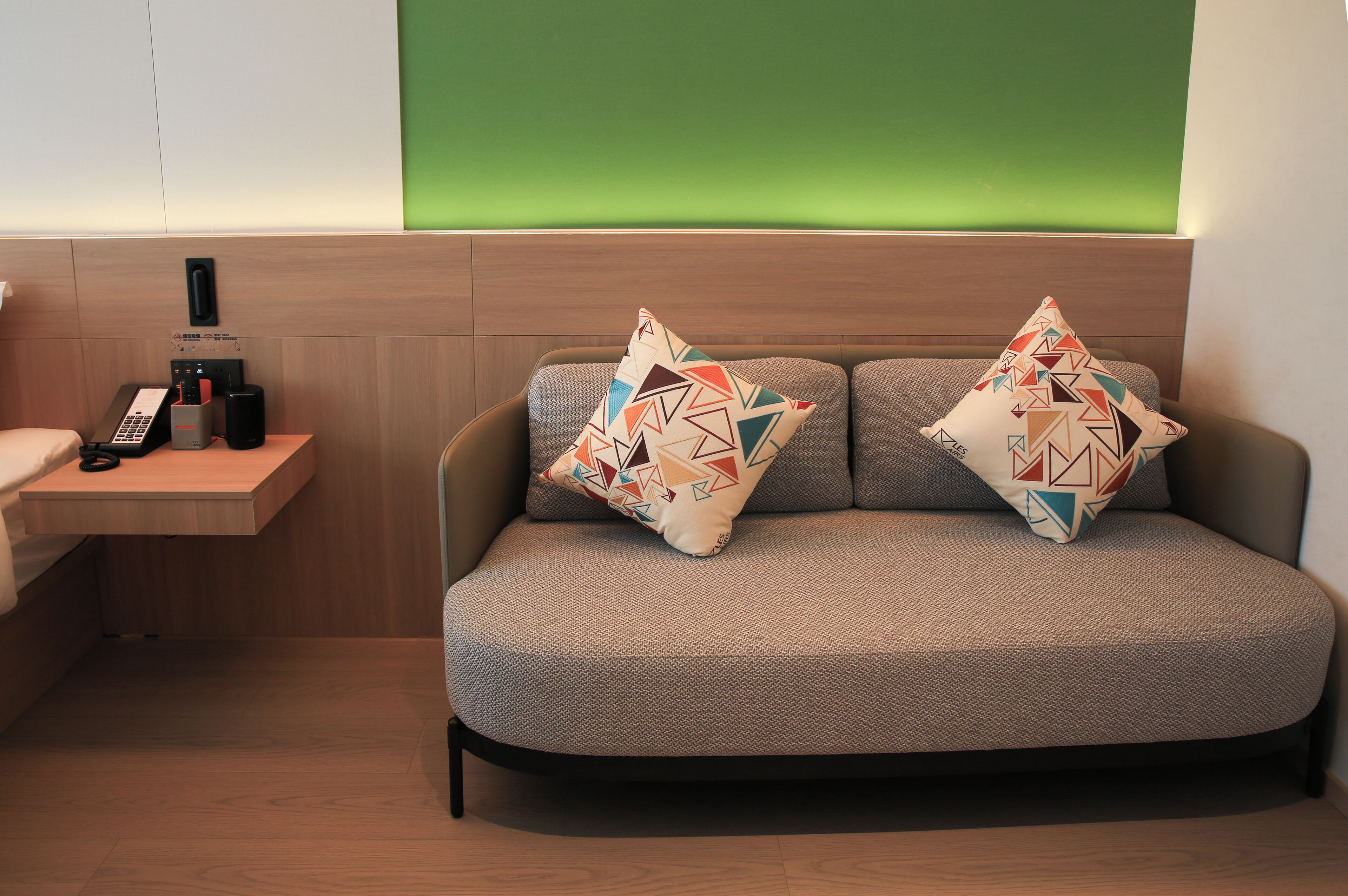 Quarto de hotel moderno com sofá, duas almofadas estampadas, mesa de cabeceira com telefone e luminária, e parede verde decorativa.