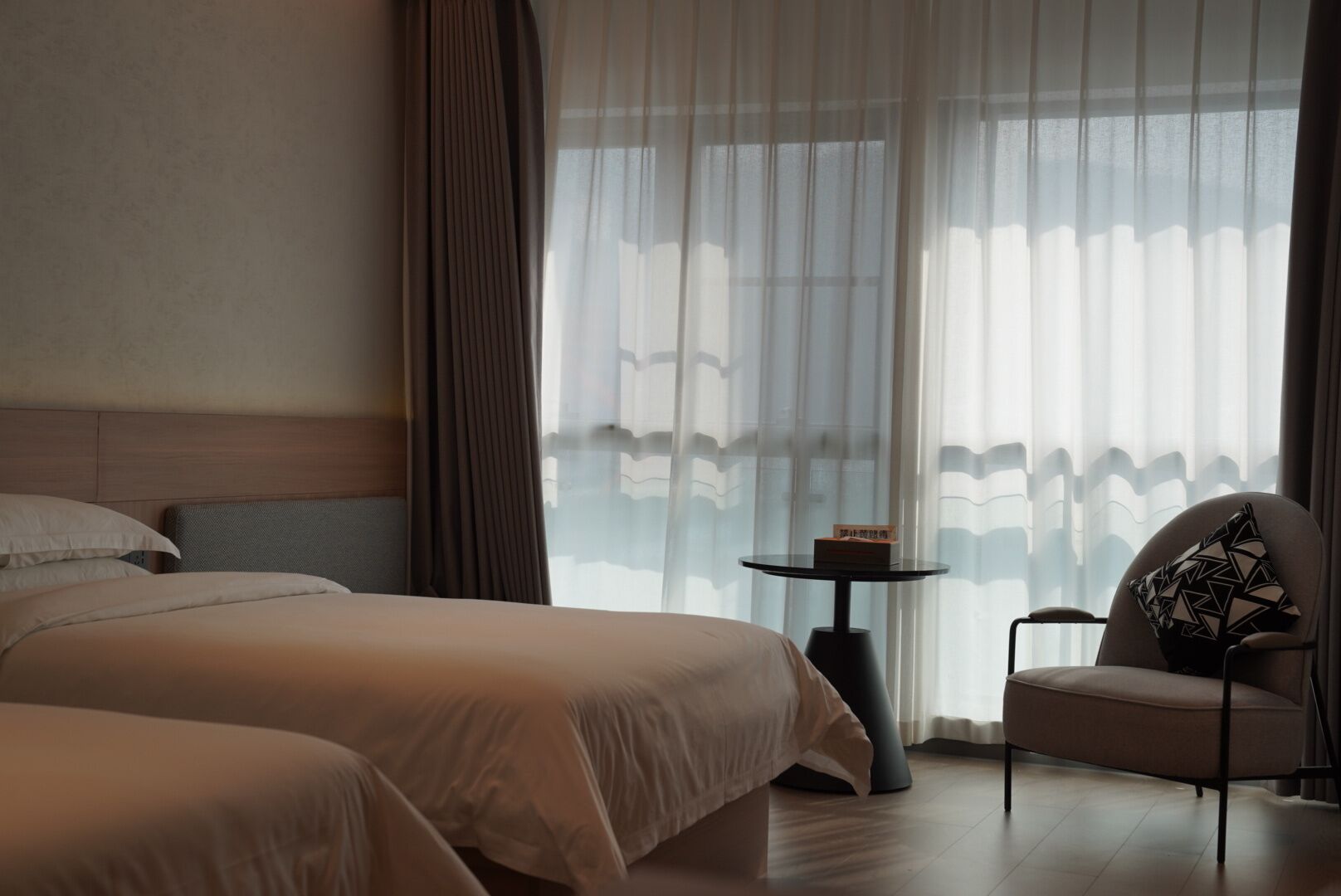 Une chambre d’hôtel avec deux lits, une petite table, une chaise et des rideaux légers.