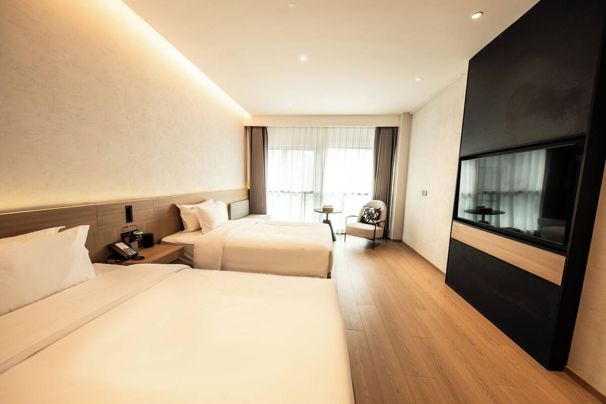 Ein modernes Hotelzimmer mit zwei Betten, einem kleinen Tisch, einem Sessel, einem Fernseher und einem Fenster mit Vorhängen.