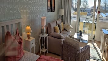 Ein gut beleuchteter Raum mit einem Bett, einer Couch und einem Korbstuhl. Ein Fenster bietet Blick auf die Gebäude vor dem Haus.