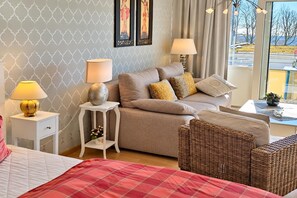 A bedroom with a bed, two bedside tables with lamps, a sofa, a wicker chair, and a window with a view.