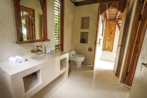 A bathroom with a white sink, a toilet, and a mirror. There are towels on the sink and a wooden door leading to another room. - Pelangi Tree House at Inspirit Ubud (Bali)