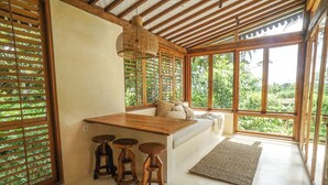 A room with a wooden ceiling, large windows, a wooden table with stools, and a view of greenery outside.