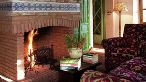 A brick fireplace with a fire, a potted plant, and books on a table. - El Águila, Stone Cottage (Alájar)