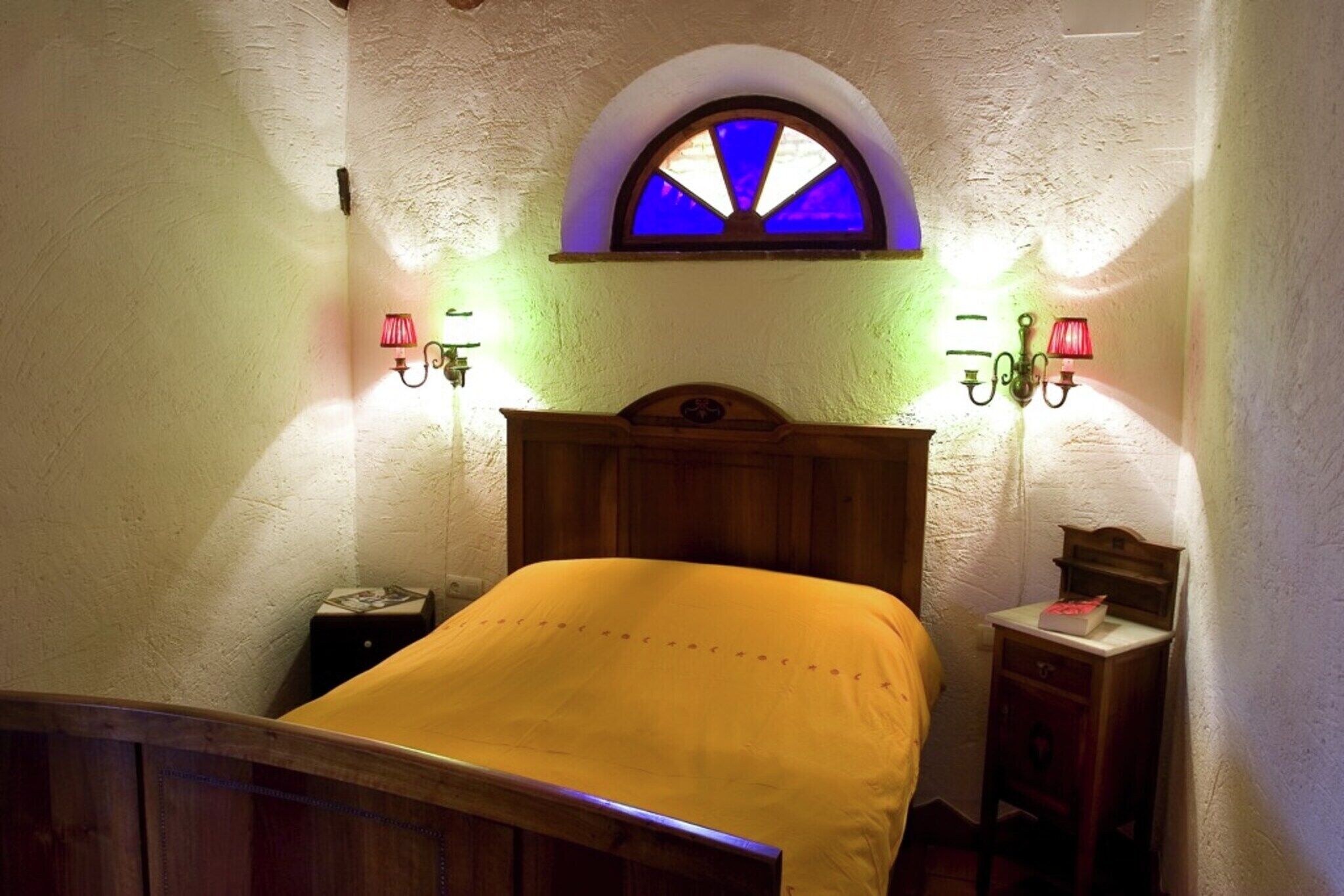A room with a bed, a wooden headboard, a small bedside table, a nightstand, a wall-mounted shelf, and a window with a stained glass design.