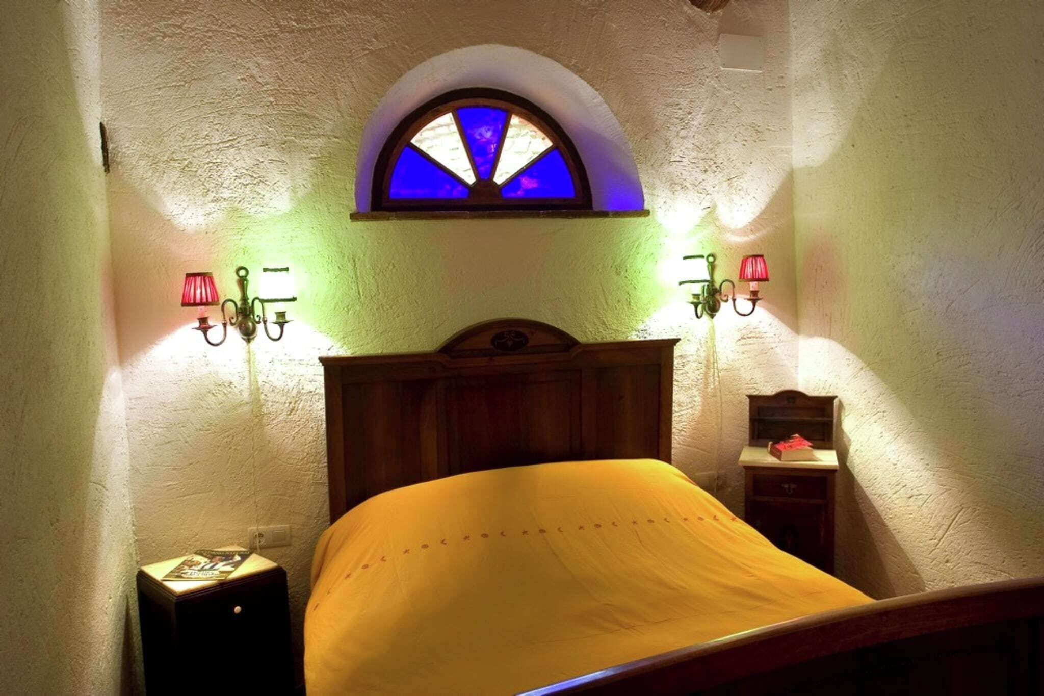 Een slaapkamer met een houten hoofdbord, een bed met een gele sprei, een klein nachtkastje met een lamp en een wandplank.