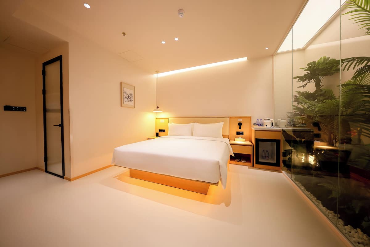 A modern bedroom with a large bed, bedside tables, a nightstand, a framed picture, and a glass enclosure with plants.