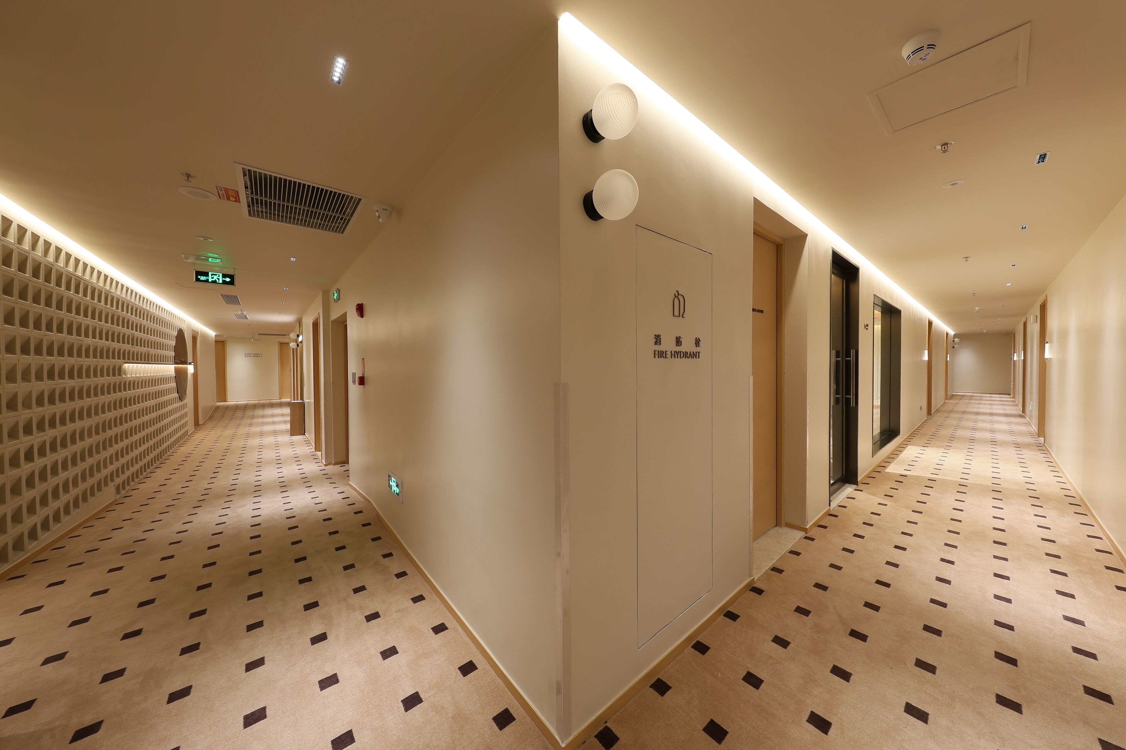 A hotel corridor with patterned carpet, recessed lighting, and a fire hydrant sign.