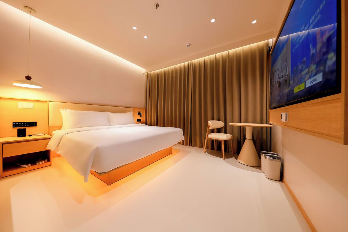 A modern hotel room with a large bed, bedside table, lamp, TV, and a small table with a chair.