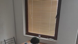 Een klein zitehoekje met een tafel, twee stoelen, een raam met zonwering, een potplant en een placemat.