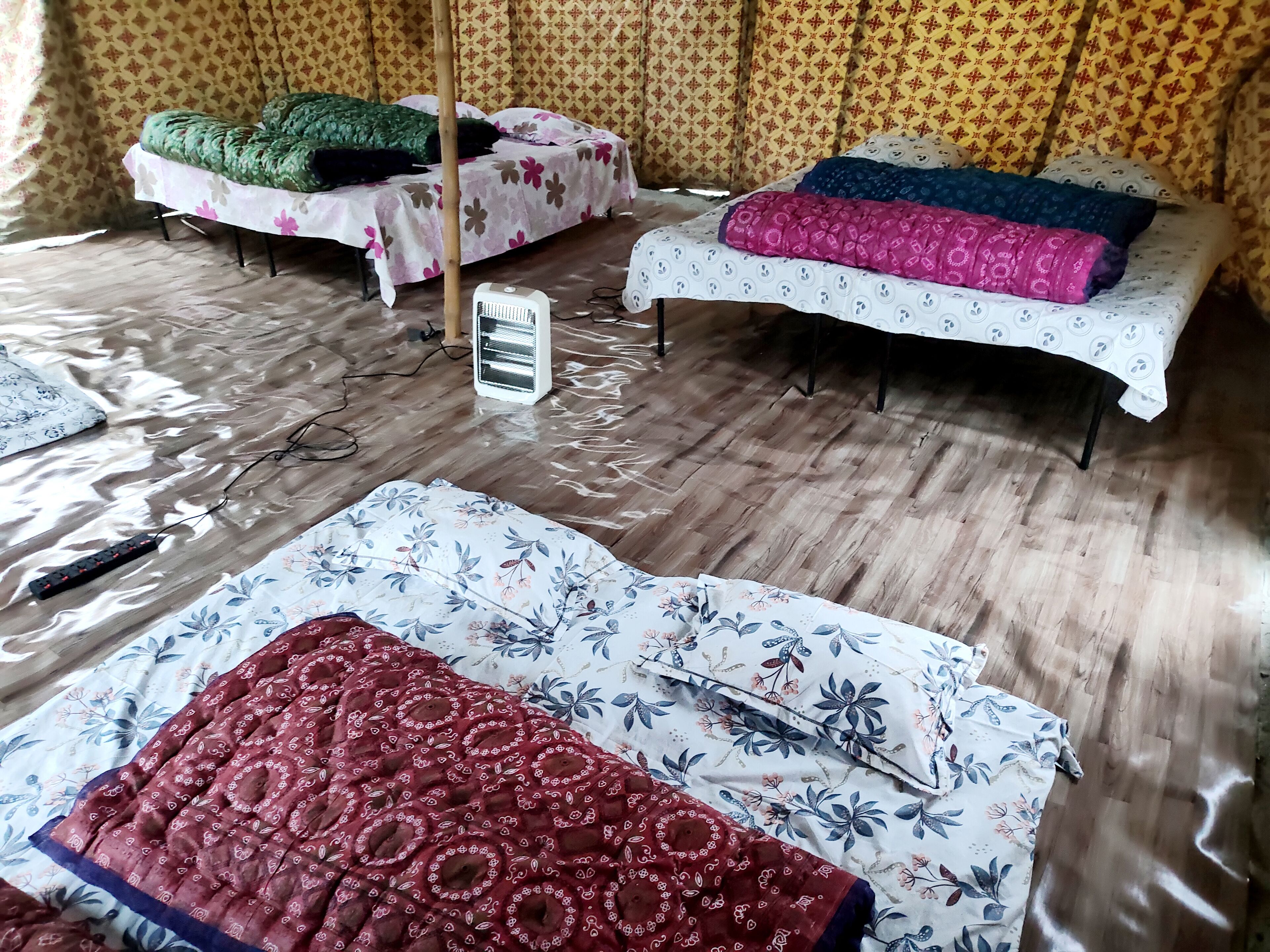 A room with two beds, one with a green blanket and the other with a pink and blue blanket. There is a heater on the floor and a power strip with a phone charger.