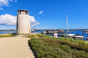 A waterfront walkway leading to a cylindrical structure with a viewing platform, a marina with boats, and a clear blue sky.