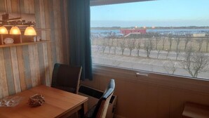 A room with a wooden table, chairs, a window with a view of a distant building and trees. - Holiday Home in Fehmarn near Beach (Fehmarn)