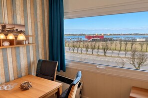 A room with a wooden table, chairs, a window with a view of a body of water and industrial structures, and a curtain.