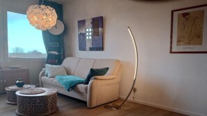 A living room with a sofa, a round coffee table, a floor lamp, and a window with a view. - Holiday Home in Fehmarn near Beach (Fehmarn)
