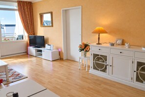 A modern living room with a flat-screen TV, a small table with a lamp, a cabinet with glass doors, and a window with curtains. - Holiday apartment with terrace in Fehmarn (Fehmarn)