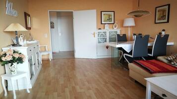 Ein Wohnzimmer mit Essbereich, Holzboden, einem weißen Sofa, einem Couchtisch, einem Fernseher und einer Küchenzeile mit Schränken und einem Kühlschrank.