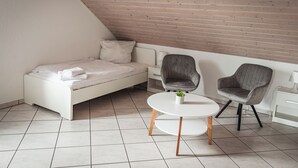 A modern bedroom with a white bed, two grey armchairs, a small white table, and a wooden sloped ceiling.
