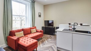 Ett modernt vardagsrum med en röd soffa och ett tillhörande soffbord, en platt-TV och en köksdel med vita skåp och en svart bänkskiva.