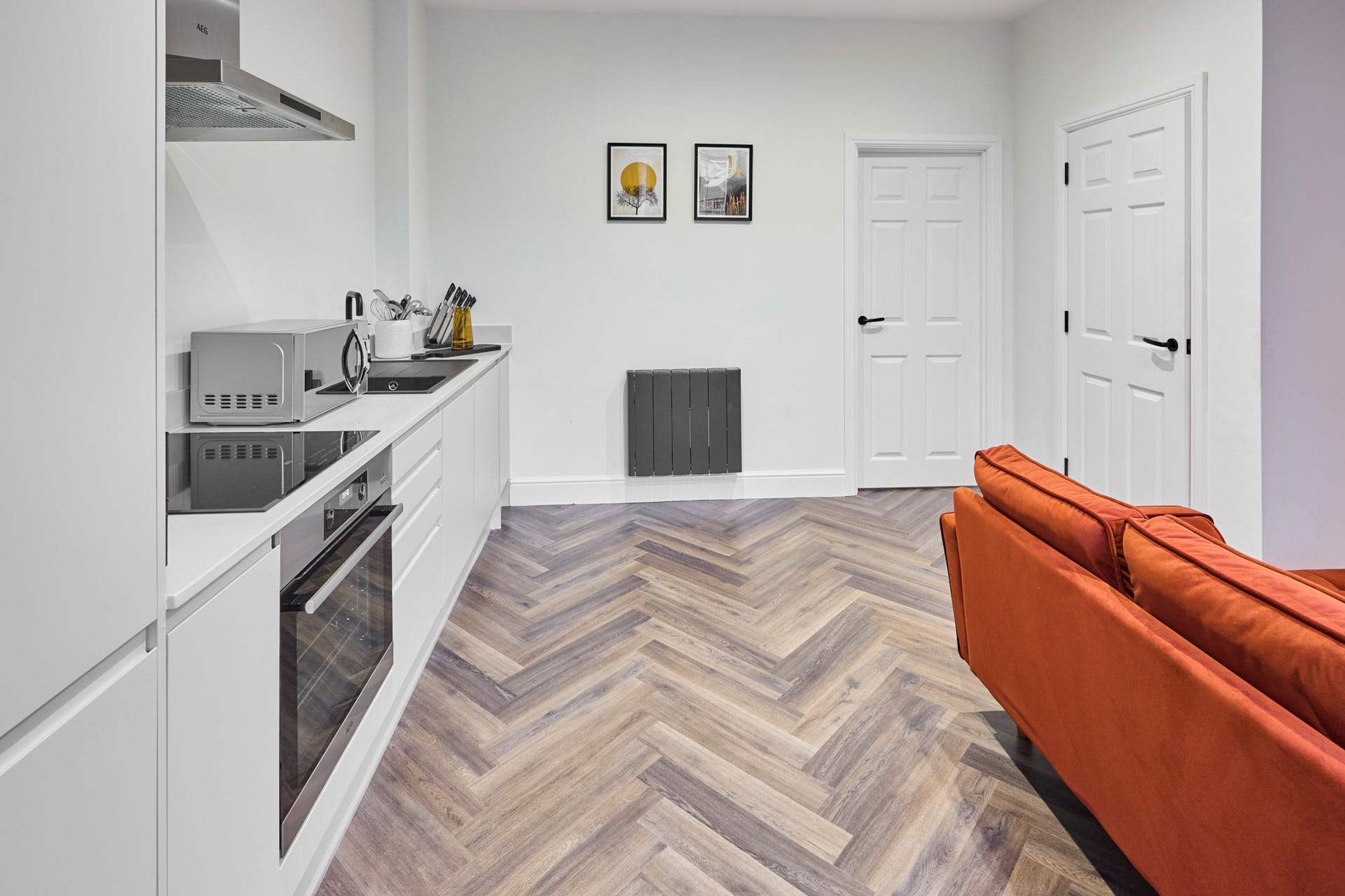 A modern kitchen with white cabinetry, a built-in oven, and a microwave. There is a herringbone-patterned wooden floor and an orange sofa in the living area. Two framed pictures are hung on the wall.