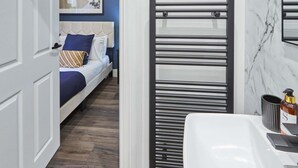A modern bathroom with a white sink, a black towel rack, and a bed with blue bedding in the adjacent room. - Host Stay Apartment 1 Upton House (Northampton)