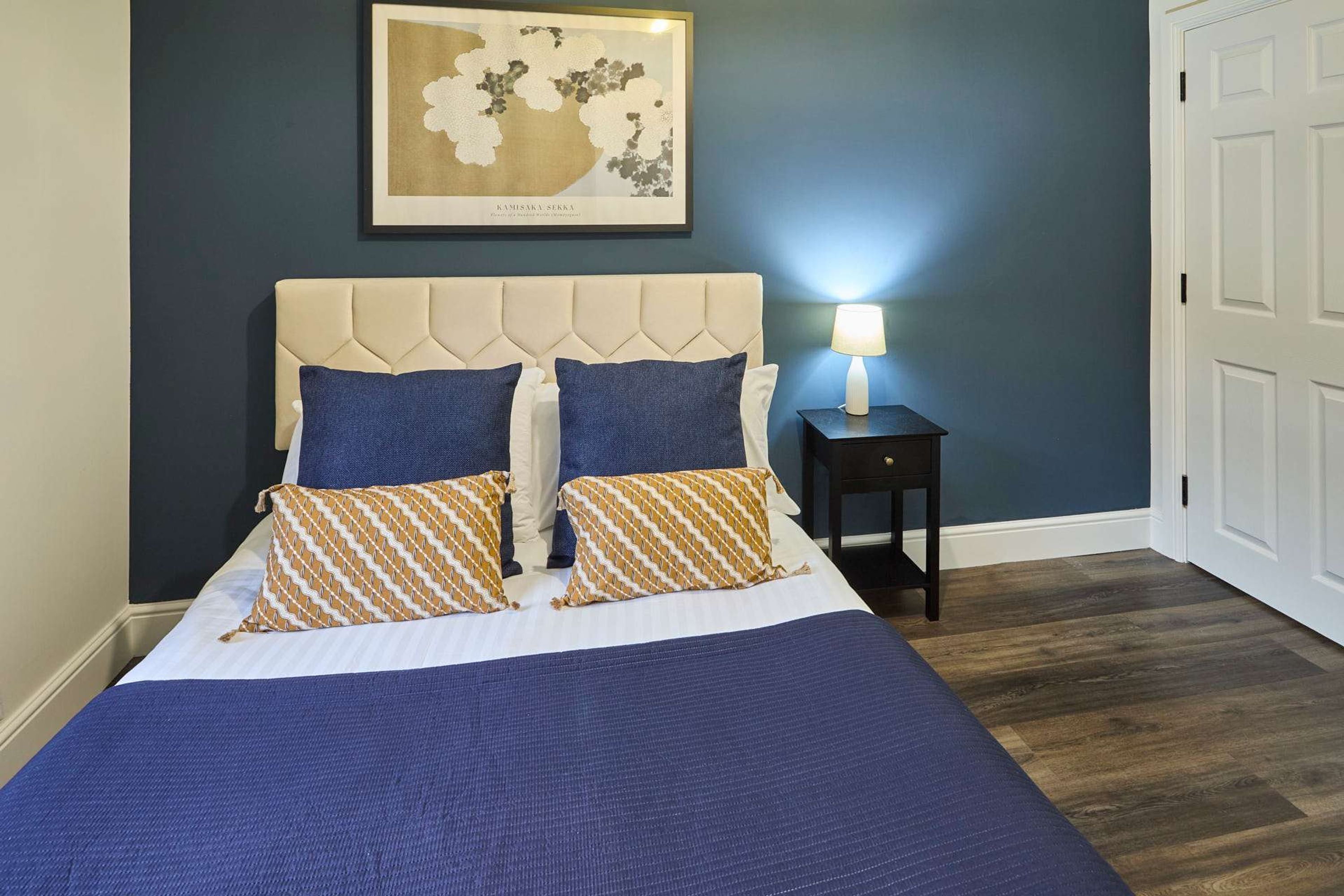 Een net opgemaakt bed met een blauw-witte spreideken, twee blauwe kussens en twee kussens met een patroon. Boven het bed hangt een ingelijste kaart aan de muur.