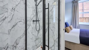 A bathroom with a marble wall, a shower with a black frame, and a bed with a blue blanket and pillows.
