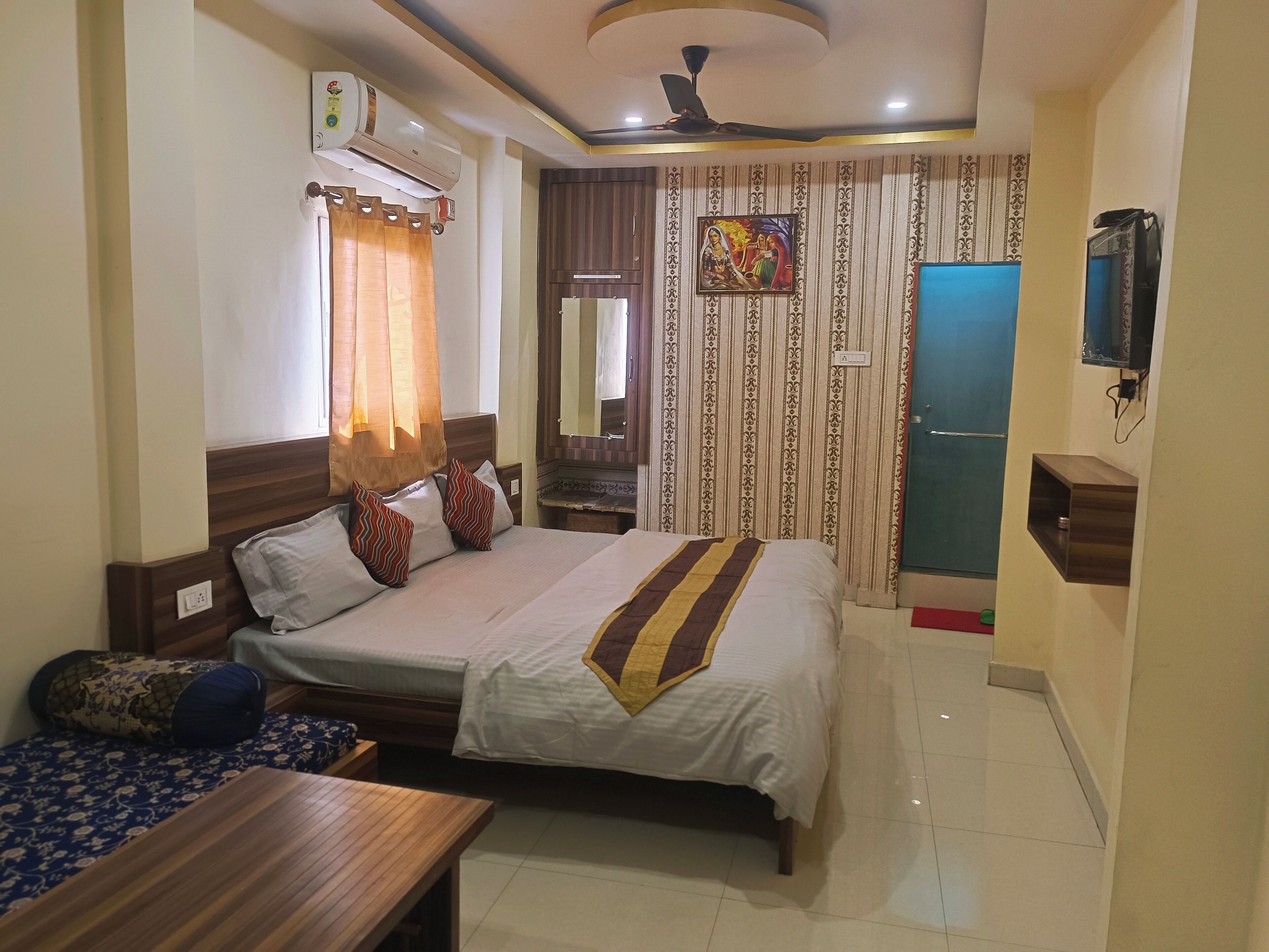 A hotel room with a bed, a wooden nightstand, a small table, a ceiling fan, a TV, and a bathroom door.