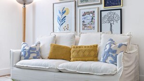 Un canapé blanc agrémenté de coussins à motifs, un lampadaire et des œuvres d’art encadrées accrochées au mur.