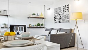 Een moderne woonkamer met een grijze bank, een witte salontafel en een eethoek met een witte stoel en borden. Er zijn planken met decoratieve objecten en een vloerlamp.
