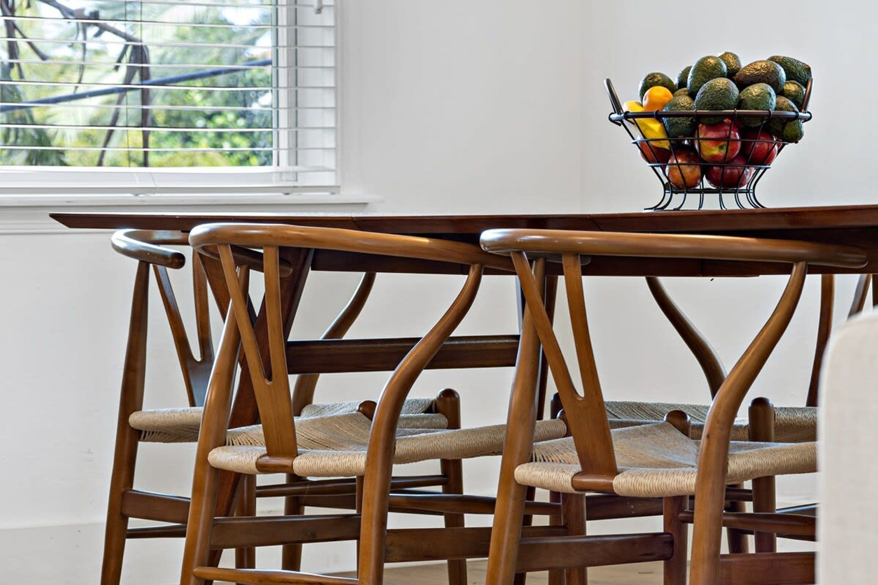 A dining area with a wooden table and chairs, a fruit basket on the table, and a window with blinds.