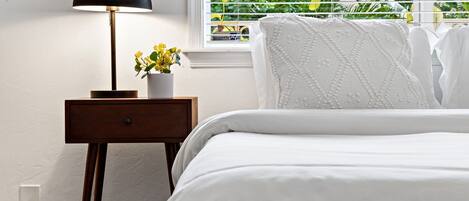 Una cama bien hecha con ropa de cama blanca, una mesita de noche con una lámpara y una ventana con persianas.