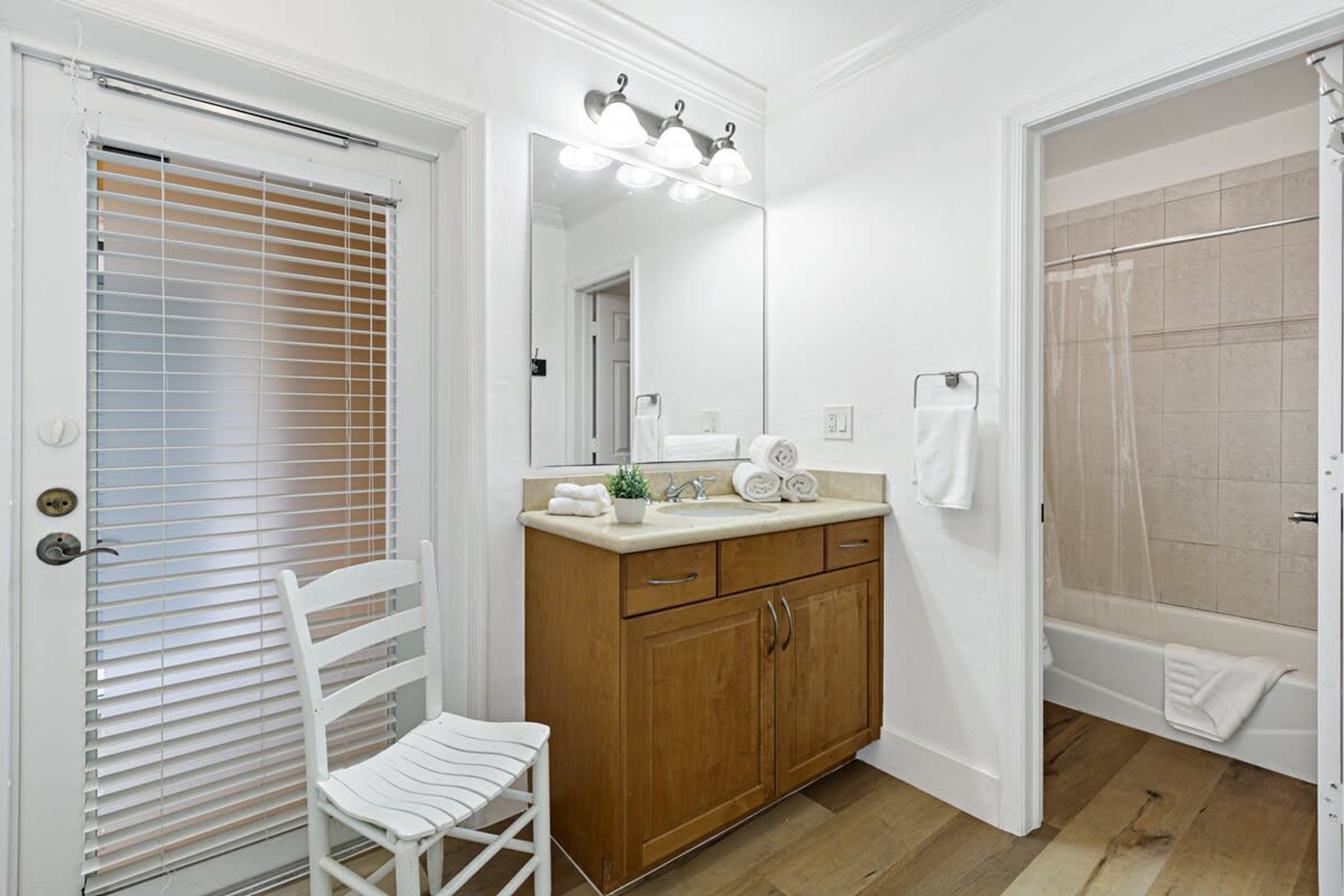 Une salle de bain avec un meuble-lavabo en bois, une chaise blanche, un miroir avec éclairage intégré, une douche et une porte avec des stores.