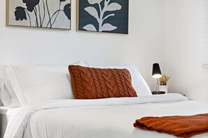 A neatly made bed with white linens and a brown knitted throw, a bedside table with a lamp, and two framed floral artworks on the wall. - Lovely Rental Unit With Free Parking on Premises (Fort Lauderdale)