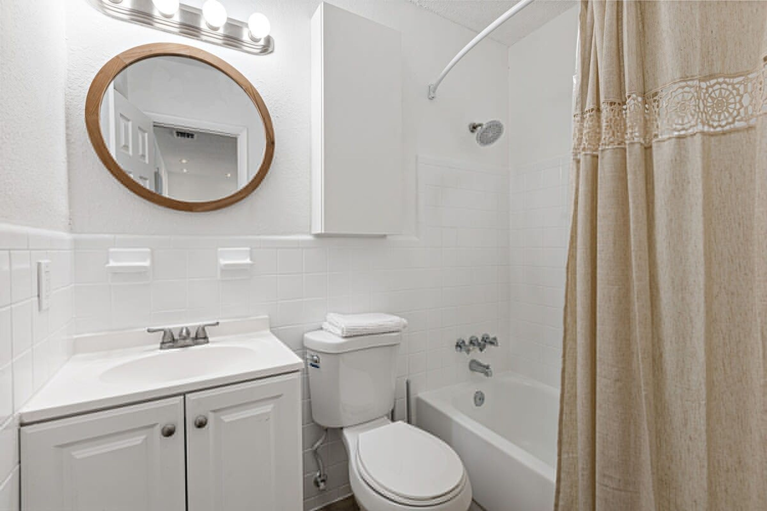 Une salle de bain avec un lavabo, des toilettes et une baignoire, tous de couleur blanche. Un miroir avec des lumières est placé au-dessus du lavabo, et il y a un rideau de douche.