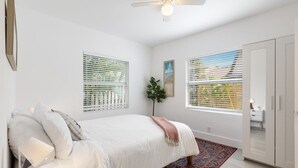 Une chambre à coucher comprenant un lit, un ventilateur de plafond, deux fenêtres équipées de stores, une plante et une armoire.