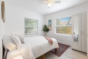 Een slaapkamer met een bed, een plafondventilator, twee ramen met zonwering, een plant en een kledingkast.
