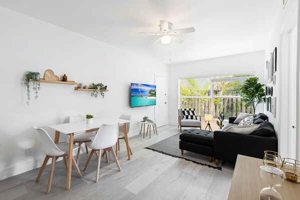 A modern living room with a black sofa, a dining area with white chairs, and a TV mounted on the wall.