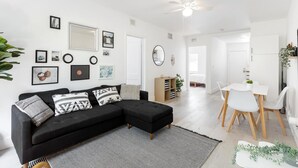 Un salon moderne avec un canapé d’angle noir, un coin repas avec des chaises blanches et un ventilateur de plafond.