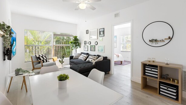 Un salon moderne avec un ventilateur de plafond blanc, un canapé noir agrémenté de coussins à motifs, une table à manger blanche et un buffet en bois doté de boîtes de rangement.