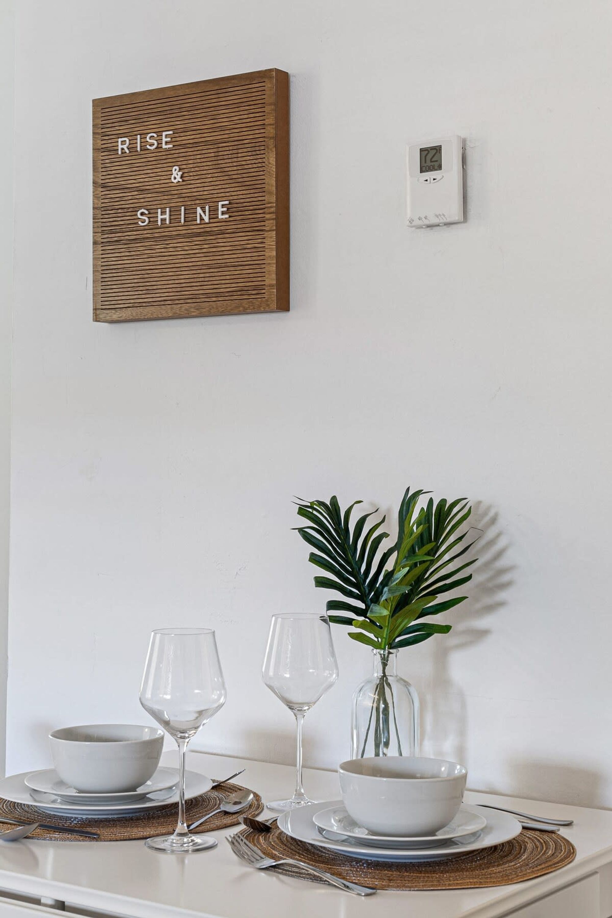 A dining table with a white tablecloth, two wine glasses, a bowl, and a plant. A wooden sign with the words "RISE & SHINE" is mounted on the wall above the table.