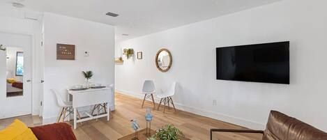 A modern living room with a brown leather sofa, a wooden coffee table with a plant, a dining area with a white table and chairs, and a mounted TV on the wall.