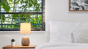 Una cama bien hecha con ropa de cama blanca, una mesita de noche con una lámpara, una planta pequeña en maceta y una ventana con persianas.