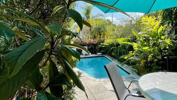 Une piscine entourée d’une végétation luxuriante, avec un espace salon abrité et un grand parasol offrant de l’ombre.